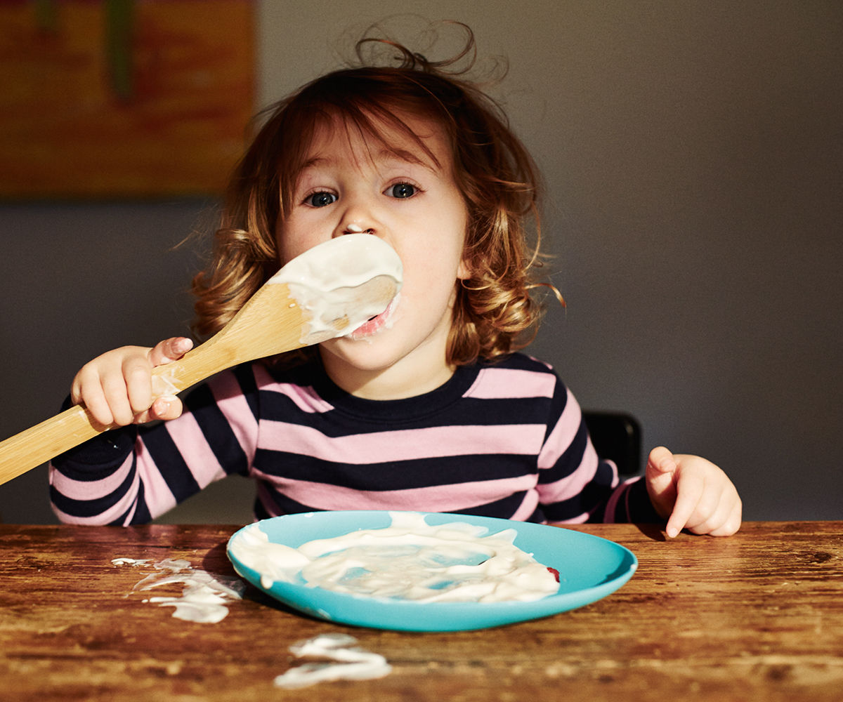 A young child licking yoghurt from a wooden spoon